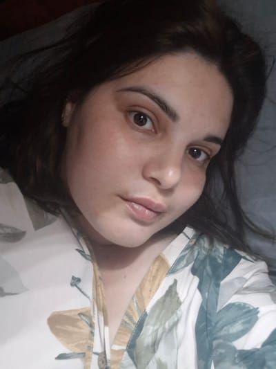 A woman with dark hair is lying down, her head tilted towards the left. She has fair skin and is wearing a patterned top with floral designs in shades of white, yellow, and blue. The background appears to be a pillow or bedding in a solid grey color. The photo seems to be taken indoors. There is no visible indication of the location being in the city of Амзя, Russia.