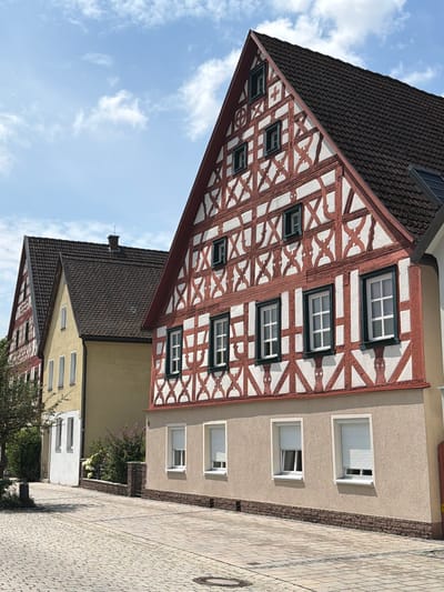The image shows a view of traditional German timber-framed houses in Ebermannstadt, Germany. The house in the foreground features a prominent red and white timber-frame facade with multiple windows. To the left of this house, another timber-framed house is partially visible, followed by a yellow building with white trim. The street in front of the houses is paved with cobblestones, and there is a stone wall with some vegetation behind it. The sky is blue with scattered white clouds, indicating a daytime scene. There are no people visible in the image.