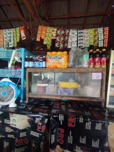 This image depicts a close-up view of a small, rustic storefront or warung, characteristic of a local convenience stall in Kecamatan Arjasa, Indonesia. No people are visible.
The scene is dominated by various goods displayed for sale. Rows of individually packaged sachets, likely containing instant drinks or snacks, hang from strings across the upper portion of the stall, with some clearly showing the brand "ZINC" on white packets. Below these, on a wooden shelf, a multipack of "Floridina" orange-flavored drinks is prominently featured. To the right of the Floridina, several bottles filled with a dark liquid are arranged, alongside clear plastic bottles. On the left, a light blue plastic shelving unit holds additional items, and a small, light blue and white electric fan sits on a surface, suggesting the need for cooling.
A glass-fronted display case with a wooden frame is situated beneath the main shelf, containing various small, indistinct items. The foreground is occupied by large, black bags, possibly containing bulk goods like coffee or snacks, which are stacked on a counter. These bags are emblazoned with large white and red "PRO" branding. Visible text on these bags includes motivational phrases in Indonesian such as "1% BAKAT 99% KERJA KERAS NEVER QUIT," "GAGAL ITU BIASA TANTANGAN DATANG BUT NEVER QUIT," and "SAATNYA UNTUK BERAKSI NEVER QUIT." One bag also mentions "COFFEE BREAK." The ceiling above is constructed from corrugated metal supported by wooden beams, indicating a simple, sheltered structure. The overall impression is a bustling local shop offering a variety of everyday items.