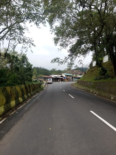 The image depicts a two-lane asphalt road in Bangtsho, Bhutan, curving gradually to the right and receding into the mid-ground. The road surface appears damp and features a dashed white center line and a solid white line marking the right edge. On the left side of the road, a low concrete barrier, painted with alternating yellow and dark green diagonal stripes, runs parallel to the road, separating it from a dense line of trees and foliage. Beyond this barrier and trees, a cluster of single-story structures with varied roof types, likely commercial establishments or residences, lines the road. A white utility vehicle or small bus with a blue stripe is parked on the left shoulder near these buildings, facing away from the camera. One individual is visible standing near the front driver's side of this vehicle.
On the right side of the road, a concrete curb and a stone retaining wall support a steep, grassy embankment. White rectangular structures or sections are visible along this embankment, suggesting further retaining walls or small buildings set back from the road. Large, mature trees with dense green foliage overhang the road from both sides, forming a natural archway at the top of the frame. In the background, beyond the immediate structures, additional hills covered in lush green vegetation rise, partially obscured by a light atmospheric haze under an overcast, light grey sky. A utility pole with a streetlamp is discernible on the right side, near the bend in the road. A blue scooter or motorcycle is partially visible parked further down on the right side of the road, near the buildings.