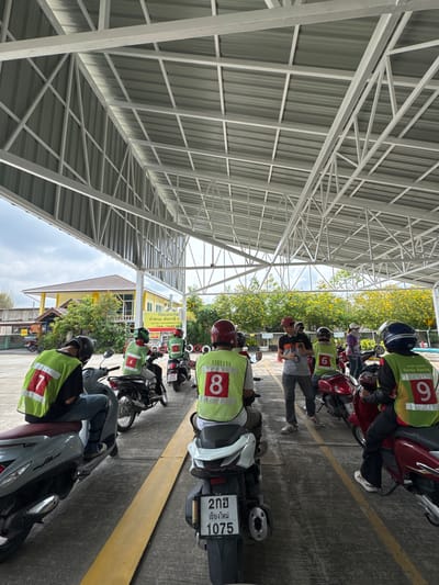 The image depicts a motorcycle parking area, likely in Nong Phueng, Thailand. Several motorcycles are parked in a row under a metal roof structure. Riders wear green vests with numbers on them, likely indicating a training course. One bike has the license plate "2กฮ เชียงใหม่ 1075". A building with a sign is partially visible in the background with text, "วิริยะประกัน ทำพรบ. ต่อภาษีด CAR TAX CAR INSURANCE CONTA". The area seems to be near a building providing car tax and insurance services. Some yellow flowering plants are also visible in the background.