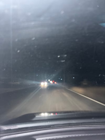 A nighttime view from a car's side mirror shows a highway receding into the distance. Bright headlights and taillights of vehicles punctuate the darkness. Streaks of light from oncoming cars create a hazy glow on the road surface. The side mirror frame is visible on the right side, with a blurred reflection on the left. The scene conveys a sense of speed and the isolation of driving at night.