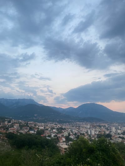 This media file depicts a panoramic view of a coastal town nestled against a mountainous backdrop. The scene is captured from an elevated vantage point, looking down upon a dense collection of buildings with red-tiled roofs and white facades, characteristic of Mediterranean architecture. Tall, modern buildings are interspersed among the smaller structures, suggesting a blend of old and new development. Lush green vegetation, including trees and shrubs, covers the foreground and spills down the slopes, adding a vibrant contrast to the urban landscape.

The sky is filled with dramatic, heavy clouds that obscure the sun, creating a somewhat moody atmosphere. The soft, diffused light suggests that it is either early morning or late evening, likely close to sunset, given the subtle warm hues visible near the horizon behind the mountains. The mountains themselves are dark and imposing, their rugged contours silhouetted against the sky.

The town appears quiet and undisturbed, with no visible people or vehicles to indicate specific activity. The overall impression is one of serene, natural beauty combined with a lived-in, established community. The location context of Šušanj, Montenegro, suggests a coastal European setting, likely in the Balkans. There is no visible text in the image.