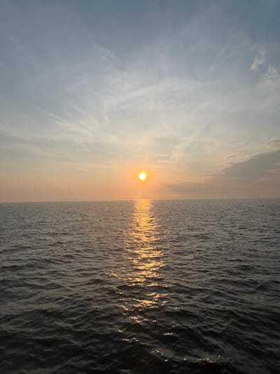 A serene sunset over the ocean with a sun reflecting on the water. The sky is a gradient of soft oranges and blues, with wispy clouds. The ocean is dark and choppy, with a bright golden reflection of the sun stretching across the surface. The horizon is visible in the distance, appearing hazy. The scene is calm and tranquil, suggesting a peaceful evening. The location is the Saint John Harbor in Saint John, Canada.