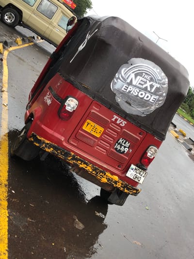 The image shows a red auto-rickshaw, commonly known as a tuk-tuk, parked on a paved street in Arusha, Tanzania, at the coordinates -3.362 latitude and 36.68 longitude. The tuk-tuk is facing away from the camera. The tuk-tuk's rear end is visible, revealing a black canvas canopy with a silver "The Next Episode" sticker. The tuk-tuk is parked on an asphalt surface with yellow painted lines and a reflection of the tuk-tuk in a puddle on the street. The rear bumper of the tuk-tuk is slightly chipped and faded with worn-out yellow paint.  There is a large "TVS" logo and a  license plate "MC 423 DHO" visible on the rear of the tuk-tuk. The tuk-tuk's license plate number is "T 751 A," and another plate reads "KLM 1449." The image is taken from a slight angle, revealing part of a tan safari vehicle parked in the background.  The scene is likely taken in a parking lot or a street corner with a wet asphalt surface.