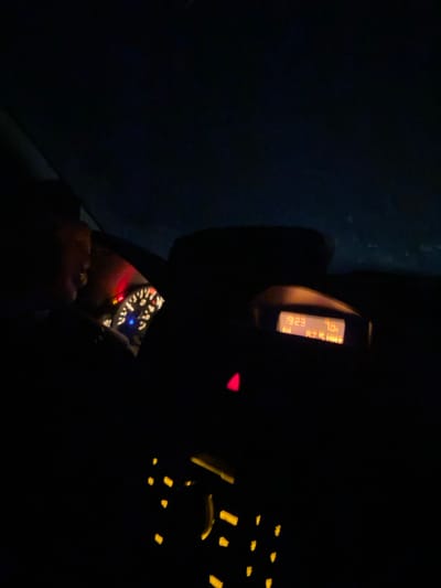 The image depicts the interior of a vehicle at night, observed from a front passenger perspective. A driver's left profile, including the head and shoulder, is visible on the far left. The dashboard is illuminated, with a visible instrument cluster displaying several lit warning indicators in red and a blue light, alongside a partially visible dial with a white needle. To the right of the instrument cluster, a digital display shows "19:23" (time), "7.0 C" (temperature), and "FM 87.5 MHz" (radio frequency). Below this display, a red triangular hazard light button is illuminated. Further down on the center console, numerous small, rectangular buttons are visible with amber backlighting, likely control elements for the vehicle's entertainment or climate systems. The exterior beyond the windshield is dark, indicating nighttime conditions. The specific city location of Podaiva, Bulgaria, is not discernible from the image data.