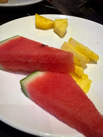 A close-up, overhead shot shows two triangular slices of watermelon and several pieces of pineapple on a white plate. The watermelon slices are bright red with a green rind. The pineapple pieces are yellow and cut into rectangular and triangular shapes. The plate is on a dark surface. The scene is likely from a restaurant or buffet in Carretera De San Martín, Spain. There is no clear indication of people or specific actions occurring.