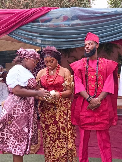 This image captures a traditional Nigerian ceremony, likely a wedding or engagement, taking place outdoors in Sabon Gari West, Nigeria. The scene features three prominently dressed individuals in the foreground, with other attendees visible in the background.

On the right, a man, possibly the groom, stands wearing a vibrant red traditional outfit that includes an embroidered tunic, trousers, and a matching cap. He has a full beard and is accessorized with multiple black beaded necklaces and bracelets. His gaze is directed slightly to his left, and his expression is serious.

In the center, a woman, presumably the bride, is elegantly dressed in a deep red traditional gown adorned with intricate gold embroidery and beadwork. She wears a prominent red beaded necklace, matching earrings, and a dark headwrap (gele). Her attention is focused downward on a small, light-colored cylindrical object she holds, indicating a ritualistic moment. Gold, leaf-like decorations embellish her arm.

To the left, another woman assists the central figure. She is dressed in a white long-sleeved top, a patterned purple and brown skirt or wrapper, and a coordinating headscarf. Her head is bowed as she participates in the action with the central woman.

The setting appears festive, with red and blue/green draped fabrics forming an overhead canopy. Green foliage from trees is visible beyond the decorations, and red carpeting covers the ground. The overall atmosphere suggests a significant cultural event, with a sense of focus and solemnity among the participants. No visible text is present in the image.