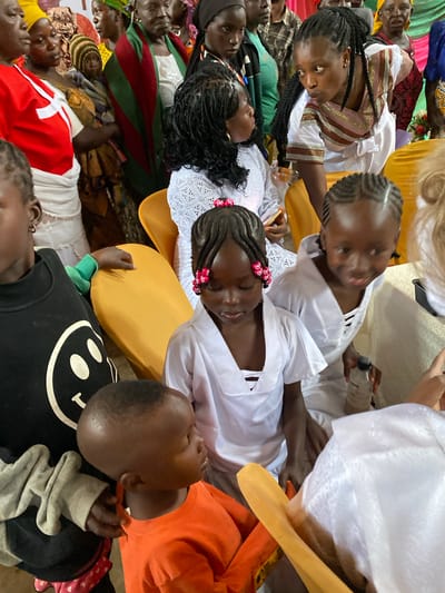 In the city of Riyom, Nigeria, a group of individuals, primarily women and children, are gathered indoors. The lighting suggests an indoor setting, possibly a community center or a large room. Several children are the focus of the image, some wearing white garments, possibly ceremonial attire, and others in casual clothing.

In the foreground, a young child in an orange shirt is being held by another person whose arm is visible. To the left, a child in a black t-shirt with a smiley face design is also visible. In the center and to the right, children in white outfits, some with braided hair adorned with pink beads, are seated or standing.

The adult women in the background are dressed in various colorful fabrics and headscarves, common in Nigerian attire. Some appear to be observing the children, while others are engaged in conversation. The overall atmosphere seems to be one of a community gathering or a special event. The presence of chairs and the general indoor setting indicate a social event.