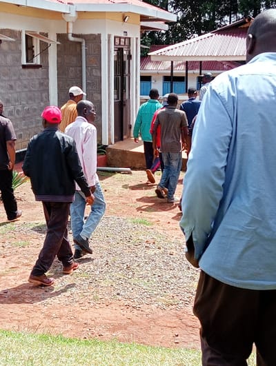 The image shows a group of seven men walking away from a building in Kipkarren, Kenya (latitude: 0.59, longitude: 34.93). The men are walking on a dirt path that is surrounded by green grass. The building appears to be made of concrete and has a red tile roof. The man in the foreground is wearing a light blue shirt and brown pants. The men are all wearing casual clothing, with one man wearing a white hat. The men are walking away from the camera, which is positioned behind them. One man is wearing a red cap. The man in the foreground appears to be blocking the view of the remaining men. The building appears to be a residential or commercial building. The scene appears to be set in a rural area. The day is sunny and there is no rain. The image does not show what the men are doing or where they are going.  The men appear to be in their mid-twenties to mid-forties. The image is blurry. The image appears to have been taken with a mobile phone.
