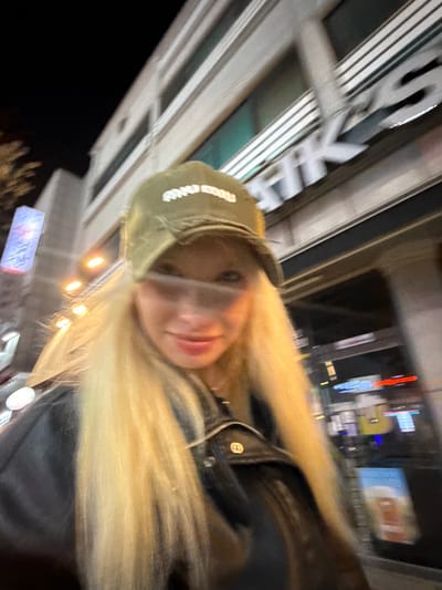 A blurry, low-angle photograph captures an individual in the foreground within an urban environment identified as Namyangju-si, South Korea, at night. The subject, a person with long, straight blonde hair, is wearing an olive green baseball cap with "miu miu" text embroidered in white, and a dark, possibly black, leather-style jacket with a visible zipper and a silver-toned button on the upper left chest. The subject's face is partially obscured by the cap's brim and horizontal motion blur, revealing light-colored eyes and a slight smile. The background features a multi-story building with a light-colored facade, displaying a large illuminated sign with partially visible white English characters (e.g., "TIKS") and Korean text. To the far left, a tall, vertical illuminated sign with blue and red elements and Korean script is visible. Further to the right, a blurred illuminated display shows what appears to be a graphic of a beer glass. The scene is illuminated by artificial light sources from streetlights and building signage.