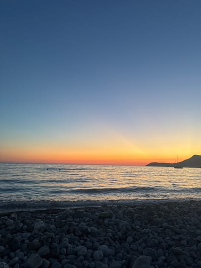 A serene, wide-angle view of a pebble beach in Šušanj, Montenegro, at dusk. The sky transitions from a deep blue at the top to a gradient of orange and yellow near the horizon, with faint rays of light fanning out. The calm sea reflects the sky's warm hues, with gentle waves lapping at the shore. In the distance, a single sailboat is visible on the water, and a silhouetted mountain range forms the backdrop on the right. The foreground is dominated by a dense collection of smooth, grey pebbles of various sizes. Two dark, circular objects, possibly buoys, are barely visible in the water, approximately one-third of the way from the left edge of the frame. The lighting suggests it is either sunrise or sunset.