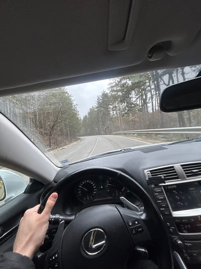 The image provides a first-person perspective from inside a moving vehicle, specifically a Lexus sedan. A light-skinned hand, presumably the driver's, is visible gripping the lower-left portion of the black, multi-function steering wheel, which prominently displays a silver Lexus emblem. The dashboard's instrument cluster indicates a speed of approximately 60 km/h and an odometer reading of 2749. Paddle shifters are visible behind the steering wheel. The center console features an infotainment system labeled "SOUND" and "CLIMATE," along with a black mounted device holder above the central air vents.

Through the front windshield, a paved two-lane road is visible, curving gently to the right in the distance. White solid and broken lane markings are present on the asphalt. A silver metallic guardrail lines the right edge of the road. Dense forest, composed primarily of tall coniferous trees and some bare deciduous trees, borders both sides of the roadway. The sky is overcast and grey, suggesting a cloudy day. The overall environment appears to be a rural or semi-rural forested area. The scene is located in Sarantsi, Bulgaria.