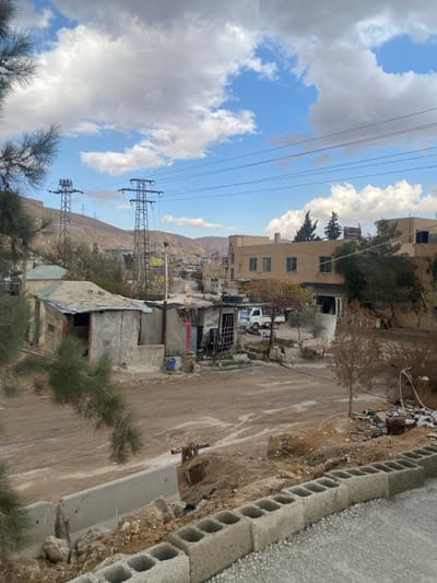 The image depicts a semi-urban landscape in Dahiyat Harasta, Syria, under a sky with significant white cumulus clouds against a clear blue background. The foreground shows a low retaining wall constructed from hollow concrete blocks, with a partially visible broken concrete barrier and loose dirt behind it. Pine tree branches extend into the left side of the frame. The midground features a wide, unpaved dirt road or open area with tire tracks. To the left of this road are several makeshift structures constructed from concrete blocks and corrugated metal, with one exhibiting a blue bucket nearby and another a red door. To the right, a multi-story, light-colored building with multiple windows stands alongside a white pickup truck parked on the unpaved surface. A pile of debris is visible on the far right of the unpaved area. The background reveals a hilly, arid terrain with two prominent metal lattice electricity pylons supporting power lines that crisscross the sky. Additional buildings and a utility pole with a yellow sign are visible further down the road, leading into the distant settlement. No individuals, explicit actions, or interactions are visible within the frame.
