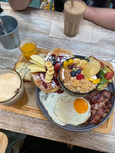 A top-down view shows a substantial breakfast platter served on a wooden board, arranged on a whitewashed wooden table. The platter contains an array of food items, including: pancakes topped with powdered sugar, fruit (banana slices, berries), and a reddish sauce; a bowl containing yogurt, granola, peanut butter, and various fruits like grapes, blueberries, raspberries, and mango; avocado toast garnished with tomato and leafy greens; grilled bacon; and a sunny-side-up fried egg. Accompanying the platter is a small glass of orange juice, a tall glass containing a light brown smoothie or shake with a straw, and a metal container filled with utensils. Another similar glass with drink is on the corner. A person's arm is visible in the top left corner. The overall setting suggests a breakfast or brunch service at a cafe or restaurant. While there is no information to suggest the specific location in Oviedo/Uviéu, Spain.