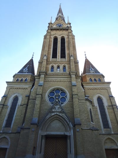 This is a low-angle, full-shot photograph of a large, brick church, likely taken on a clear day with bright, natural light. The church has a tall, central spire with a clock face near the top, adorned with Roman numerals. Flanking the central structure are two smaller, pointed spires, each topped with a cross. The church is constructed of yellow bricks and features several arched windows, including a prominent rose window above the main entrance. Decorative stonework, including pointed arches and finials, embellishes the facade. Two tall, narrow windows are visible on each side of the central section. The main entrance is a large, wooden door with a patterned design, set within a grand arched entryway. The sky above is a clear, pale blue, suggesting it is daytime, possibly late afternoon due to the lighting. No people or specific activities are depicted, and no visible text is present within the image itself, though the Roman numerals on the clock are a form of text. The architectural style suggests a Gothic Revival or similar European ecclesiastical design. The image appears to be a documentation of the church's exterior architecture.