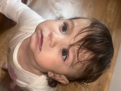 A close-up, upward-facing shot depicts a young child with light skin, brown hair, and brown eyes. The child is wearing a light-colored, long-sleeved shirt. The background appears to be a wooden floor. There is no discernible information connecting the image to Elena, Bulgaria.