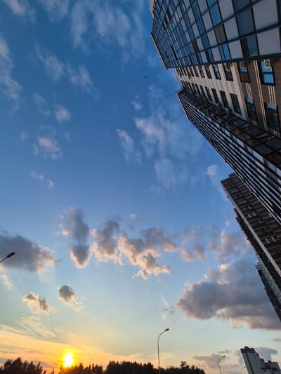 This media file depicts an upward-facing view of modern residential buildings and a sky with clouds at sunset. The buildings are tall, multi-story structures with reflective glass facades and visible windows and air conditioning units. The sky is a gradient of blue and orange, with scattered clouds, some of which are illuminated by the setting sun. The sun itself is visible on the right side of the frame, partially obscured by trees and emitting warm, golden light. A streetlight is visible in the lower right corner. Several birds are also visible flying in the sky between the buildings. The scene suggests an urban setting in Voronezh, Russia, during the evening, with a peaceful and serene atmosphere. There is no visible text in the image.