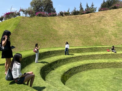 The image displays an outdoor scene at El Nido de Quetzalcóatl in Naucalpan de Juárez, Mexico, under a clear blue sky. Five individuals, appearing to be women, are situated on a multi-tiered, concave grass landscape resembling an amphitheater.

In the foreground, a woman with long dark hair, wearing a white long-sleeved shirt and blue denim shorts, sits on a lower grass terrace. She holds a dark smartphone in her left hand. To her left and slightly behind, a second woman with long dark hair, dressed in a black sleeveless dress and carrying a black shoulder bag, stands holding a dark smartphone. Adjacent to her, on a higher grass terrace, a third woman in white pants with a patterned shoulder bag holds a smartphone vertically, positioned as if capturing a photograph or video. Further to the right, on an elevated grass step, a fourth woman with long dark hair, wearing a white long-sleeved shirt and dark jeans, stands facing the camera with sunglasses on. On the far right, a fifth person, dressed in dark clothing, is seated on the highest visible grass terrace, with a bright red object on the grass beside them.

The environment consists of vibrant green, manicured grass terraces that form distinct steps. Behind these terraces, the grass slopes upwards, appearing slightly drier and browner. The background features a hilltop adorned with a variety of green trees and dense clusters of pink/purple flowering shrubs, with several conical evergreen trees visible on the right. Strong sunlight casts sharp shadows across the terraced landscape.