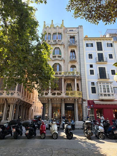 This image captures a street scene in Palma, Spain, with a prominent building in the center displaying ornate architecture, featuring arched windows, balconies, and decorative stonework. To the right, a lighter blue building with balconies and shutters stands next to it. In front of the buildings, a row of parked motorcycles and scooters occupies the paved street. A large tree with green leaves partially obscures the view of the buildings, casting shadows on the scene. The sky is clear and blue. People are visible on the sidewalk, walking or standing near the buildings. One person is standing in the doorway of a shop with the name "sessùn" visible. The overall impression is a sunny day in an urban environment with historical architecture.