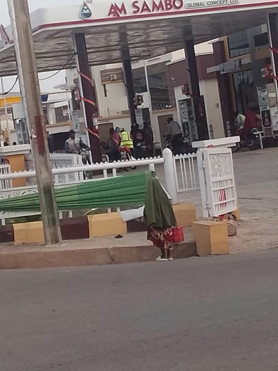 The image depicts a street scene in front of a gas station in Pantame, Nigeria. The gas station, branded with "AM SAMBO GLOBAL CONCEPT LTD.", has a red and white canopy supported by dark poles. Behind the canopy, buildings with brown and white facades are visible.
In the foreground, a person wearing a dark green headscarf and a patterned dress is standing on a raised concrete platform. They are carrying a red bag. Two yellow concrete blocks are placed to the right of this individual. A green tarp is partially visible stretched across a white fence in the mid-ground.
Behind the gas station canopy, several individuals are present, some on motorcycles. One person is wearing a bright yellow reflective vest. A white fence with decorative elements runs horizontally across the mid-ground, separating the area near the gas station from the street. A light pole is present on the left side of the image. The road in the foreground is asphalt. The overall scene appears to be a typical urban environment in Nigeria.