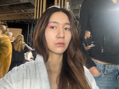 The image presents a close-up of a young Asian woman in a backstage or preparation area in Issy-les-Moulineaux, France. She has long, wavy dark brown hair and is wearing a white terrycloth bathrobe. Her left eye (viewer's right) exhibits prominent pink eyeshadow, while her right eye (viewer's left) has minimal or no visible eyeshadow. Her lips are slightly parted and glossy.

In the mid-ground to the left, a person with long light-colored hair is seen from behind, wearing a brown fur-like garment over their shoulder. Several other indistinct figures are gathered further back, appearing to be observing or waiting. Structural elements, including dark metal scaffolding and staging equipment, are visible in the background, indicative of an event setting. To the right, the torso of a person wearing a black hooded jacket and light blue jeans is partially visible, with a hand in a pocket. Behind this figure, a man with short dark hair and glasses is seen in profile, looking to the left, holding a white rectangular object. The lighting is artificial and uneven, with the foreground subject well-illuminated.