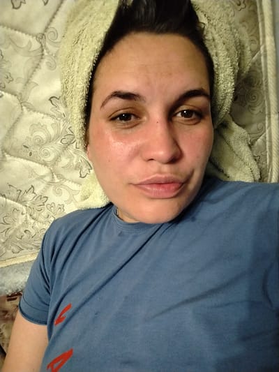 The image is a close-up, head-and-shoulders selfie of a woman with dark hair wrapped in a light-colored towel. She is wearing a blue t-shirt and appears to be lying down, with a patterned surface behind her that looks like a mattress or bedding. Her face is slightly flushed, and her eyes are open, looking towards the camera. The lighting is somewhat dim, and there is a slight sheen on her skin. There are no discernible landmarks or environmental cues to suggest a specific location within the city of Зипуново, Russia. The focus is entirely on the woman and her immediate surroundings.