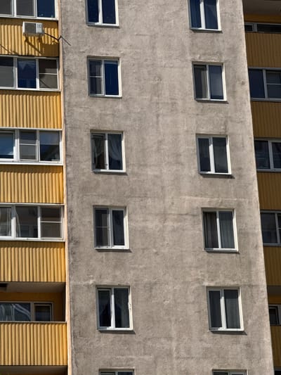 This image shows the exterior of a building with multiple stories and windows. The main subject is the building itself, constructed with a rough, gray concrete facade on the upper floors and a yellow, corrugated metal siding on the lower floors. The building features numerous rectangular windows, many of which are divided into panes by white frames. Some windows have dark glass, reflecting the surroundings, while others appear to be open or partially obscured.

On the lower level, visible to the left of the image, there is an air conditioning unit mounted on the wall, connected to the building by a thin metal pipe. Further to the right on the lower level, there is an open doorway or alcove revealing more windows and the yellow siding.

The scene is likely taking place during the daytime, as indicated by the shadows cast on the building's facade. The lighting suggests a clear day. There are no people or animals visible in the image. The location cues provided (Shushary, Russia) add context to the setting, suggesting an urban or suburban environment typical of a residential or commercial building. There is no visible text in the image.