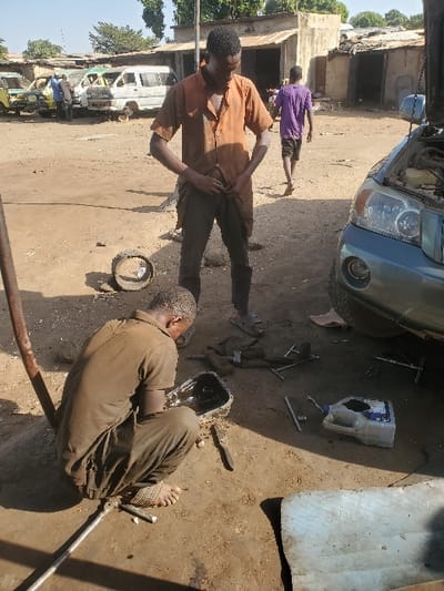 This image depicts an outdoor automotive repair scene in Kaduna South, Nigeria. The ground is unpaved earth. In the immediate foreground, a male individual in a dark brown, short-sleeved uniform squats barefoot, appearing to examine or work on a black metal pan filled with a dark liquid, possibly oil. Various hand tools, including wrenches and a long metal bar, are scattered around him. Centrally positioned, another male stands upright, wearing an orange short-sleeved button-up shirt and dark trousers, with his hands adjusting his lower front clothing. He wears sandals. To the right of the standing man, the front section of a light blue SUV is visible, its hood open, indicating ongoing maintenance. A white plastic fluid container and additional tools lie near the vehicle. In the mid-ground, several derelict white and yellow vans or minibuses are parked. In the background, a third male in a purple t-shirt walks away from the camera towards a cluster of single-story structures with corrugated metal roofs. The sky is bright and clear, and sparse vegetation is visible in the distance.