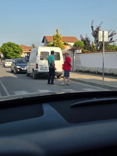 The image depicts a street scene in Yagodovo, Bulgaria, captured from within a vehicle. The foreground shows the dark dashboard of the observer's car. The midground features an asphalt road with visible pedestrian crossing markings. Two adult males are interacting on the right side of the road, adjacent to a white Ford Transit van (license plate PB 6228 TT) which is parked or stopped. The man on the left wears a light blue t-shirt, dark trousers, a dark cap, and a shoulder bag. The man on the right wears a red polo shirt, blue shorts, and black-and-white striped slides, with his hands clasped behind his back. Behind the van, a dark blue sedan (license plate PB 8...) and a white compact car are also present. A paved sidewalk runs along the right side of the road, bordered by a low white wall with decorative stone elements and terracotta tile coping. In the background, residential buildings with red-tiled roofs are visible amidst various trees and bushes, including a prominent bright green deciduous tree on the left and a yellow evergreen tree on the right. A light pole with a blank rectangular sign stands further down the street. The sky is clear and blue, indicating daytime conditions.