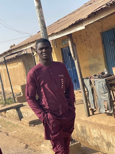 A young adult male of dark complexion, wearing a maroon traditional two-piece outfit with white chevron embroidery on the left chest pocket, stands centrally in an outdoor setting in Unguwar Gabas, Nigeria. He faces the camera directly, hands in his pockets. A utility pole is situated behind his head. The background features a faded yellow single-story building with a corrugated metal roof and blue metal doors/shutters. To the subject's right, a wooden table displays various items including sandals and a water bottle, with corrugated metal sheets leaning beneath it. The ground consists of dirt with scattered debris, and concrete steps or a raised curb are visible in the foreground. The scene is brightly illuminated by daylight.
