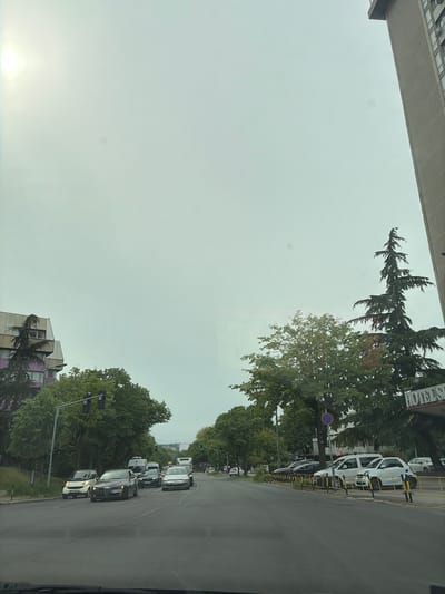 The image depicts an urban or suburban street scene under an overcast sky, with a bright spot in the lower left corner possibly indicating the sun or a strong reflection. The perspective appears to be from inside a vehicle, looking towards a road lined with trees and buildings.

On the far left, a large, modern building structure with a concrete overhang is prominently visible. The right side of the image features a multi-lane asphalt road with cars. Several white vehicles are parked along the right curb, separated from the main road by yellow barriers. Behind these parked cars, a building sign partially reads "HOTELS," framed by lush green trees, including tall conifer-like varieties and broader deciduous trees. A blue circular road sign, likely indicating "no entry" or "forbidden," is also visible near the "HOTELS" sign.

Further down the road, a line of cars is stopped in traffic. Notable vehicles in the queue include a black Alfa Romeo with the license plate "BG 2712-KX" and a white Smart car. Another vehicle immediately behind the Alfa Romeo has the license plate "BG 0244-GX." A green traffic light is visible in the distance, indicating that the traffic is likely waiting for the signal to change. The road is bordered by dense foliage, and another building with distinct purple, pink, and white sections can be seen in the distance on the left side of the road.

The "BG" prefix on the license plates confirms the location as Belgrade, Serbia, consistent with the provided context of Vozdovac Urban Municipality. The scene captures a typical daytime urban environment with traffic and mixed-use buildings.