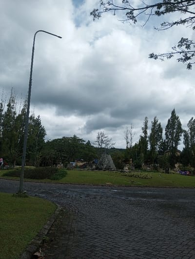 A grey brick road leads to a grassy island with a pyramid-shaped stone structure. The island is surrounded by trees of varying heights. A tall street light stands to the left of the road, extending into a cloudy sky. Branches from another tree are visible in the upper right corner of the image. The scene appears to be in a park or garden setting.