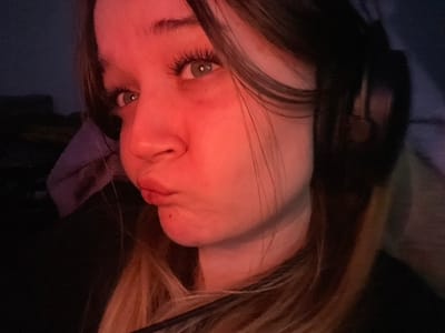 A young female subject is depicted indoors in the city of Pushchino, Russia. Her face, captured in a close-up, is strongly illuminated by a red light source from the left side of the frame, casting a reddish hue across her skin. She has long, dark hair with lighter strands visible at the bottom right. Her visible left eye is green and features prominent eyelashes. Her lips are pursed in a pout, and she maintains a direct gaze towards the camera. Dark over-ear headphones are worn on the right side of her head, partially visible. A small dark mark is present on her chin. The background is dark, out of focus, and indistinct, with a light purple fabric item visible on the far left.
