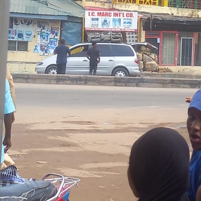 The image, taken in Sardauna, Nigeria, depicts a street scene. A white minivan is parked along the side of the road. Two men are standing near the van. One is wearing a dark jacket and the other a blue shirt. A sign for "I.C. MARC INT'L CO." is visible on a building. Posters are plastered on a wall near the minivan. In the foreground, parts of people, possibly vendors, are visible with merchandise. The road is dusty. Buildings with shops are visible in the background.
