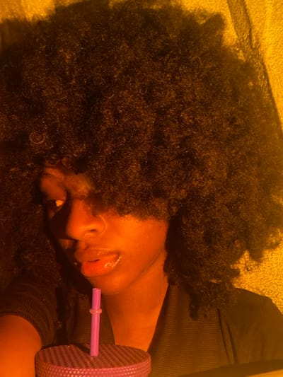 An image taken in Iranla, Nigeria, depicts a close-up, waist-up view of a young adult individual with dark skin and a voluminous Afro hairstyle. The individual is positioned slightly to the right of the frame, looking towards the left. Their left eye is partially obscured by hair and shadow, while the right eye is visible, gazing slightly upward. Their lips are parted, appearing glossy, with the lower lip positioned near a pink or purple plastic straw. The straw extends vertically from a textured, lidded cup, dark purple or pink in color, which is held or placed in the lower-left foreground. The individual wears a dark, possibly ribbed or textured, garment. The scene is illuminated by strong, warm yellow-orange light originating from the right, casting deep shadows on the left side of the face and within the hair. The background consists of a uniformly warm-toned surface.