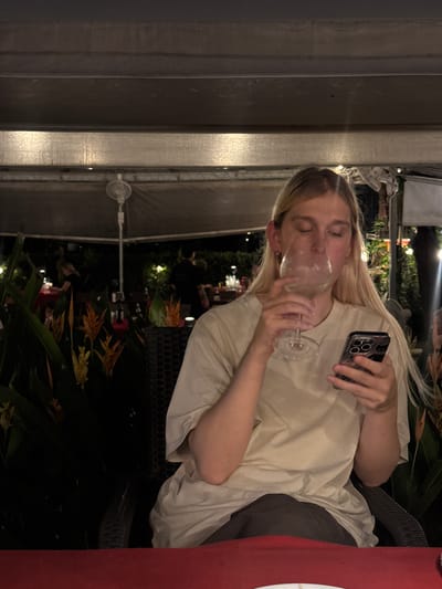 The image shows a young person with blonde hair at an outdoor dining establishment. The person is wearing a light-colored t-shirt and dark pants. They are holding a wine glass in their right hand and a smartphone in their left hand. The person is drinking from the wine glass, and their eyes are closed. The background is dark, with some artificial lights visible, suggesting it is nighttime. There are tropical plants and foliage in the background, which, along with the dim lighting and outdoor setting, hint at a warm climate. The person appears relaxed or perhaps in a moment of enjoyment. The image is oriented vertically, with the person positioned on the left side of the frame. A red tablecloth or surface is partially visible on the right side of the image. The overall impression is a casual evening out.