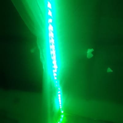 A vertical chain of bright, cyan-colored lights is set against a dark green background. The string of lights appears slightly blurred. Scattered in the green space around the light string are faint, distorted, ghostly shapes. There is a dark, undefined form on the left side of the image. The overall effect creates an eerie or otherworldly atmosphere.