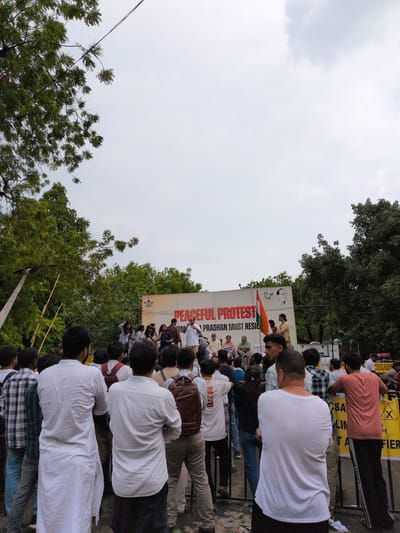 A crowd of people are gathered for a peaceful protest in New Delhi, India. The image captures a side view of the event, with a banner prominently displayed that reads "PEACEFUL PROTEST PRADHAN MUST RESIGN". An Indian flag is also visible. The sky is overcast, suggesting a cloudy day. The people appear to be predominantly male, dressed in casual attire. Some individuals are looking towards the banner, while others are engaged in conversation. The overall mood seems to be one of focused participation in the demonstration. Trees and foliage frame the scene, indicating an outdoor setting, possibly a street or public square. The presence of barriers suggests a controlled or organized gathering.
