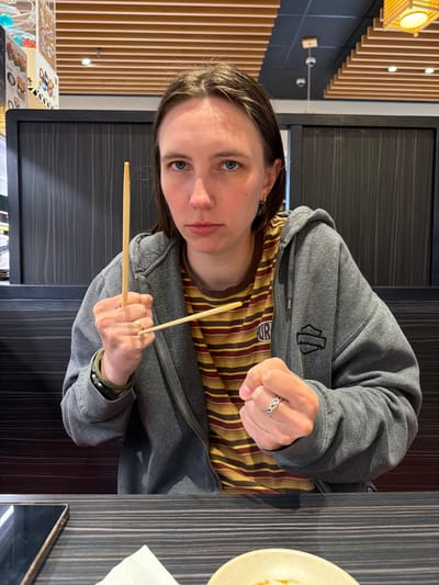 A woman with brown hair is pictured in a restaurant setting. She is wearing a grey hooded jacket over a striped t-shirt. Her hands are clenched in fists, and she is holding chopsticks in a playful or confrontational manner. One chopstick is held vertically between her knuckles, and another is held horizontally across her fingers. She is wearing a ring on her left hand. The background shows wooden panels and some decor, suggesting an Asian-themed restaurant. The table in front of her has a dark wood grain pattern. The scene is lit by overhead lights. The woman is looking directly at the camera with a serious expression. The location is a restaurant in Beaverton, Oregon, United States of America.