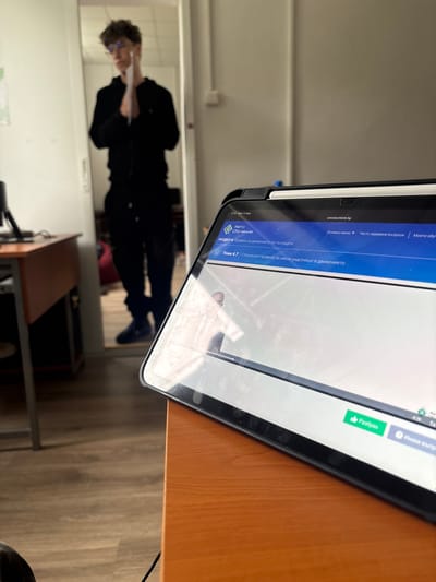 The image shows a tablet displaying a website related to driving education ("avtoobuchenie.bg"). The screen shows text in Bulgarian. In the reflection on the tablet's screen, a person in dark clothing and glasses can be seen standing in a room, possibly holding a document. The tablet is resting on a wooden desk with a cable running along the right side. The room appears to be indoors with a desk, chair and mirror. Without additional information, the location in Troyan, Bulgaria cannot be confirmed.