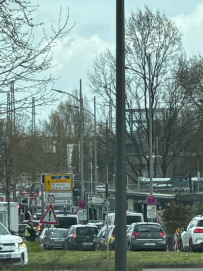 The image shows a busy street in Kehl, Germany. Multiple cars are lined up in traffic. A yellow road sign points to the left, next to a speed limit sign (30) and a triangular warning sign. A person in a yellow vest is visible near the front of the line of cars. Bare trees are visible in the background, obscuring buildings and the sky. Overhead power lines are visible above the street.