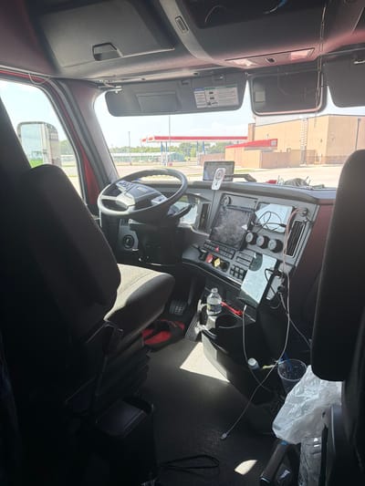 The image shows the interior of a semi-truck cabin parked outside a convenience store. The truck's dashboard is visible, featuring a steering wheel, multiple screens, and various buttons and controls. A tablet is mounted on the dashboard, and a drink cup and a plastic bag are on the passenger side. The driver's seat, a dark fabric material, is in the foreground. Through the windshield, a red-roofed building with a gas station canopy and a sign is visible, suggesting a location in Cardwell, United States. The exterior lighting indicates it is daytime. The scene is a static shot of the truck's interior and its immediate surroundings.