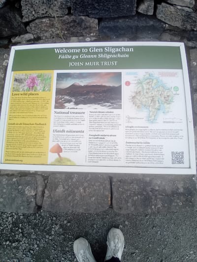 The image shows a sign at Glen Sligachan on the Isle of Skye, United Kingdom. The sign, titled "Welcome to Glen Sligachan" and "Fàilte gu Gleann Shligeachain" with "JOHN MUIR TRUST" below, is a white framed informational panel with a green border. It features text and images divided into sections. On the left, under the heading "Love wild places", there is a photograph of a pink flower and text in English and Gaelic. Below this is the website address "johnmuirtrust.org". In the center, under "National treasure", is a photograph of a mountain landscape and text in English and Gaelic, accompanied by an image of a mushroom. To the right, the top section is a map of the area with highlighted routes. Below the map, under "A fragile environment", there is text in English and Gaelic, including a QR code and the John Muir Trust logo. The sign is placed outdoors, with the base resting on a stone and gravel surface. The feet of a person wearing athletic shoes are visible at the bottom of the image, suggesting the sign is at ground level. The background is a dry stone wall.