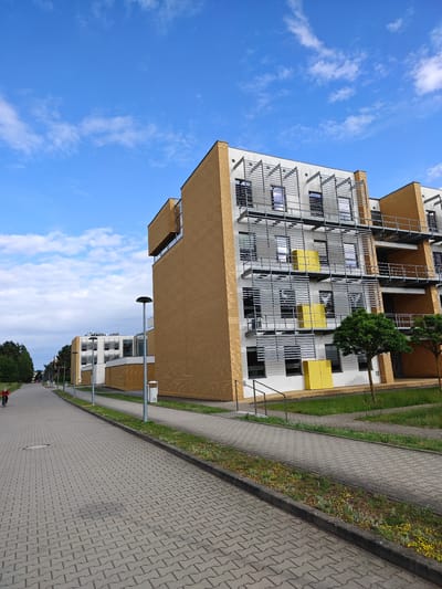 The image shows a modern apartment building on a sunny day in Poznan, Poland. The building has a yellow brick facade with white and grey accents, featuring multiple balconies with metal railings and horizontal sunshades. Bright yellow panels are visible on some of the balconies. A paved walkway runs along the building, with a grass strip and a few trees separating it from the building. In the foreground, a person on a bicycle is visible on the paved path, which leads into the distance towards more buildings and trees. The sky is blue with scattered white clouds.