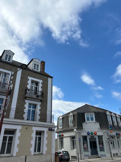 The image shows a street scene in Dinard, France. The scene is located at coordinates 48.631 latitude and -2.055 longitude, on Rue de la République. Two buildings are visible, both with multiple stories. The building on the left has white trim on the windows and beige siding. It also has a black wrought iron balcony. The building on the right has a gray roof and white trim on the windows. The building on the right has a blue awning and a sign that says "MAMA." A white car is parked in the street, near the curb. The sky is blue with some white clouds.