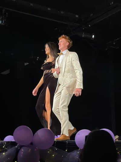 Two young adults, a female and a male, are walking side-by-side across a dimly lit indoor stage, presumably in Palos Verdes Estates, United States. The female, positioned on the left, has long dark hair and wears a dark, high-slit strapless dress with a patterned midriff section, revealing her left leg up to the mid-thigh. She has a ring on her right ring finger. The male, on the right, has red hair and wears a light-colored, patterned three-piece suit (jacket, vest, trousers), a white collared shirt, and a light-colored bow tie, complemented by brown dress shoes. Both individuals are smiling and looking towards their left (viewer's left) as they walk. The stage's edge in the foreground is decorated with several purple balloons, dark netting, and small, warm-toned string lights. The background is dark, revealing overhead stage lighting fixtures and metal rigging with numbered labels (e.g., 57, 58, 59, 60, 61). A person's head and a smartphone, held upright as if recording, are partially visible in the bottom right corner, indicating an audience present.