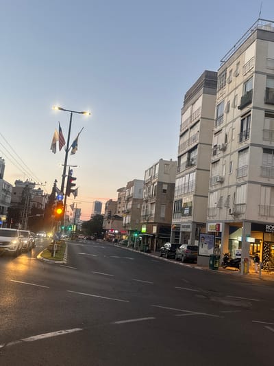 The image captures an urban street scene in Ramat Gan, Israel, during dusk or early evening. The sky transitions from a clear light blue overhead to a warm orange-pink glow near the horizon, indicating the time of day.

Multi-story residential and commercial buildings line both sides of the street. These buildings, likely constructed of concrete or stucco, feature numerous windows, many with external shutters or grilles, and visible air conditioning units. Their ground floors house various businesses with illuminated storefronts. One prominent sign reads "ROSE BAKERY." An advertising board for "FINQ" prominently displays a man's face and Hebrew text including "למה אתם מחכים?" (What are you waiting for?) and "שוק ההון בשבילכם" (The capital market for you).

The asphalt road features multiple lanes, clearly marked with white lines and directional arrows. A narrow, grassy median strip divides the road. Cars with illuminated headlights are visible, both parked along the curbs and in traffic. A motorcycle is also parked near a storefront.

Several streetlights are illuminated, casting a warm glow, and a traffic light displays an orange/yellow signal. A tall street pole holds multiple small flags, one of which resembles the American flag, along with a blue circular street sign featuring a white arrow. On the far left, a small green sign with the number "63" is visible. The scene portrays a typical urban setting during the transition from day to night.