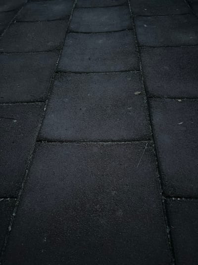The image displays a close-up, ground-level view of a paved area constructed from dark, rectangular interlocking tiles. The tiles appear to be made of a rough, granular material, possibly rubberized asphalt or a similar composite. The surface is slightly uneven, with some tiles showing subtle depressions or warping, creating a gentle, undulating texture. The lines between the tiles are visible as thin, dark grooves, with some appearing to have accumulated a small amount of debris or sediment. A few small, light-colored particles, possibly dust or small pebbles, are scattered across the surface. A single, thin, light-colored strand, potentially a blade of grass or a thin fiber, is visible lying across one of the tile seams in the lower right quadrant of the image. The lighting is dim, suggesting either an overcast day or evening conditions, and the overall color palette is dominated by dark grays and blacks. There are no people, animals, or other significant objects present in the frame. The scene is devoid of any specific context that would indicate a particular location, and no identifiable features of the city of Sinyavino, Russia, are discernible.