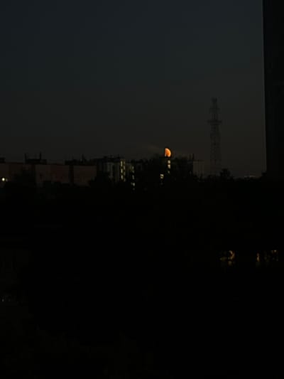 The image depicts a nighttime cityscape in Shushary, Russia, dominated by darkness and silhouettes. A crescent moon, illuminated with an orange hue, is visible in the upper portion of the frame, positioned above a cluster of buildings. A tall, lattice-like tower, likely a radio or power transmission tower, stands prominently behind the buildings, also silhouetted against the night sky. The foreground is almost entirely black, suggesting dense foliage or shadows obscuring the ground level. Scattered lights from windows in the buildings are faintly visible, indicating activity within the structures. The overall atmosphere is one of stillness and the transition from day to night.